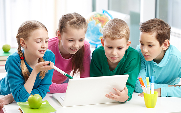 Vier Kinder stützen sich mit den Ellenbogen auf einen Tisch und blicken gemeinsam auf ein Tablet.