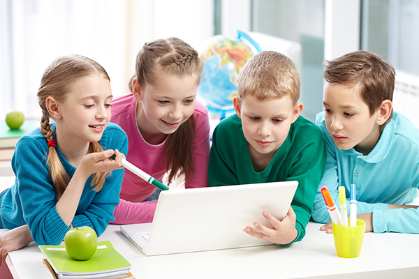 Vier Kinder stützen sich mit den Ellenbogen auf einen Tisch und blicken gemeinsam auf ein Tablet.
