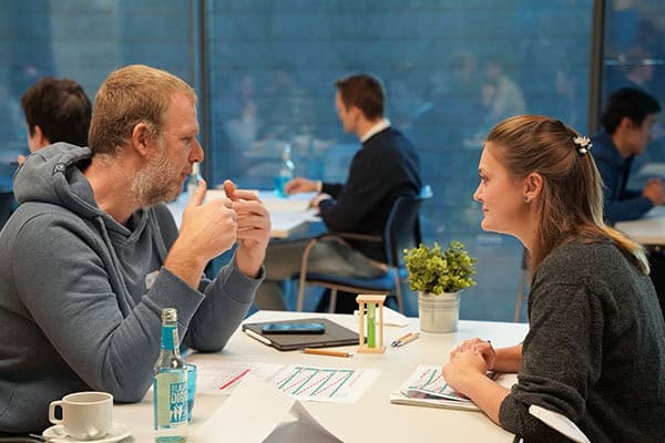 Zwei Menschen sitzen am Tisch und unterhalten sich; im Hintergrund sieht man weitere Menschen an Tischen sitzen.