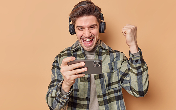 Ein Mann hat Kopfhörer auf und guckt auf ein Smartphone, dabei reißt er freudig eine geballte Faust in die Luft.