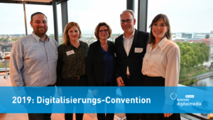 Auf dem Foto sieht man fünf Personen die nebeneinander stehen und lächelnd in die Kamera schauen. Darüber steht auf einem blauen Band: 2019: Digitalisierungs-Convention.