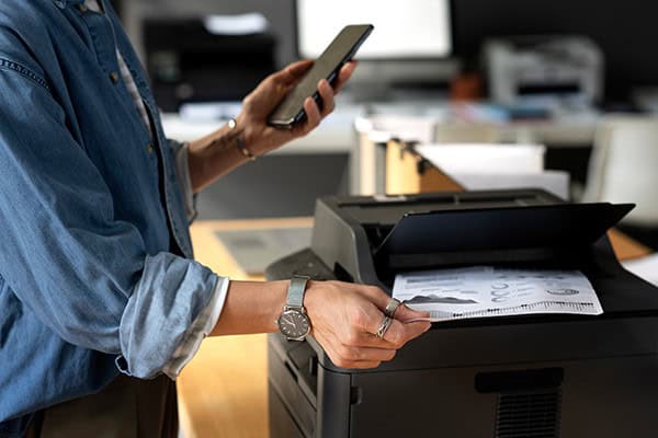 Nahaufnahme von zwei Händen: in einer ein Smartphone und die andere nimmt parallel ein bedrucktes Blatt aus dem Drucker.