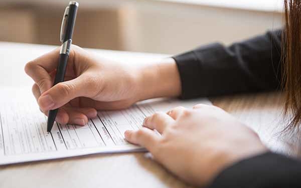 Zwei Hände liegen auf einem Tisch auf einem Blatt Papier, in einer Hand wird mit einem Stift ein Formular ausgefüllt.