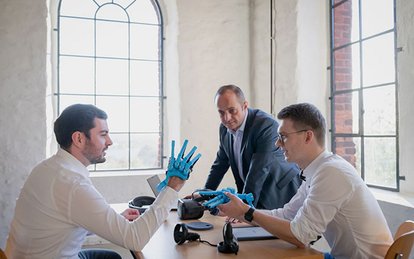 Drei Menschen sitzen und stehen um einen Tisch, einer hält eine technische Roboterprothese einer Hand in der Hand, ein zweiter hat eine weitere über die Hand gezogen.