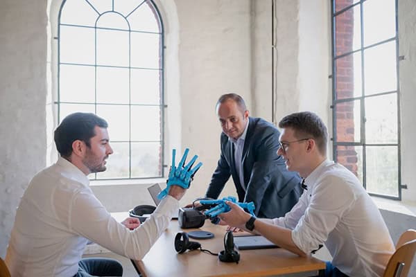 Drei Menschen sitzen und stehen um einen Tisch, einer hält eine technische Roboterprothese einer Hand in der Hand, ein zweiter hat eine weitere über die Hand gezogen.