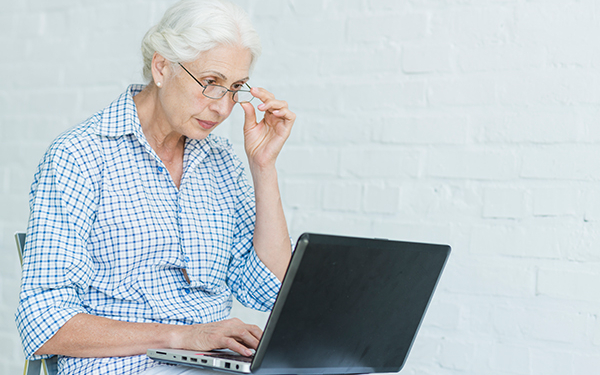 Eine Seniorin sitzt mit einem aufgeklappten Laptop auf dem Schoß auf einem Stuhl, hält mit einer Hand die Brille, die sie trägt und shaut auf den Bildschirm.