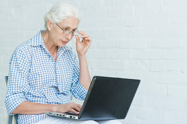 Eine Seniorin sitzt mit einem aufgeklappten Laptop auf dem Schoß auf einem Stuhl, hält mit einer Hand die Brille, die sie trägt und shaut auf den Bildschirm.
