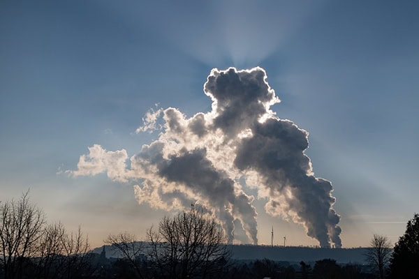 Weitsicht auf industrielle Schornsteine, die im Gegenlicht der Sonne Rauch ausstoßen.