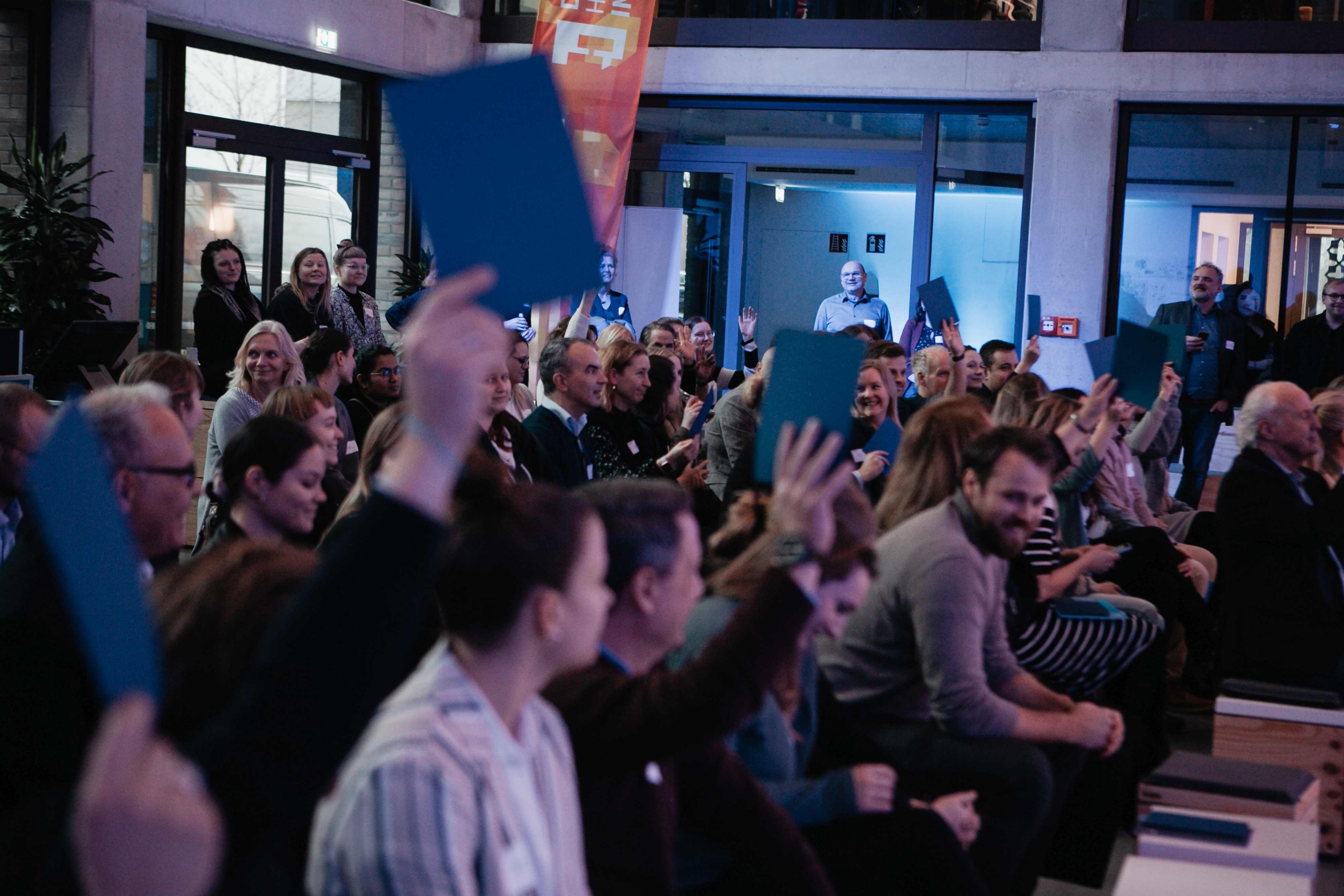 Eine große Gruppe sitzender Menschen agiert auf einer Veranstaltung mit Menschen auf der Bühne außehalb des Fotobereichs. Einige halten blaue Karten in der Hand.in die Luft.