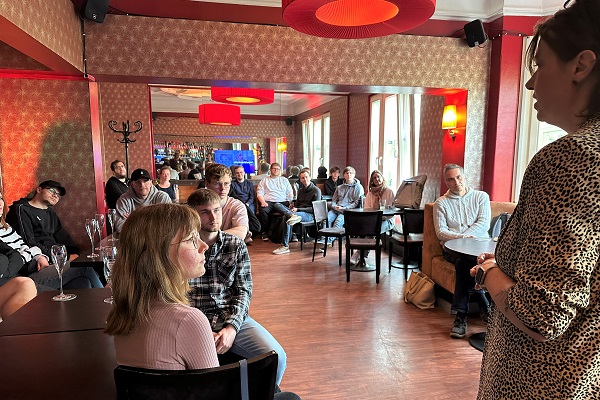 Blick in einen rötlich tapezierten Raum, indem rund 15 Personen sitzen und einer Person zuhören, die vorne (am Bildrand) steht.