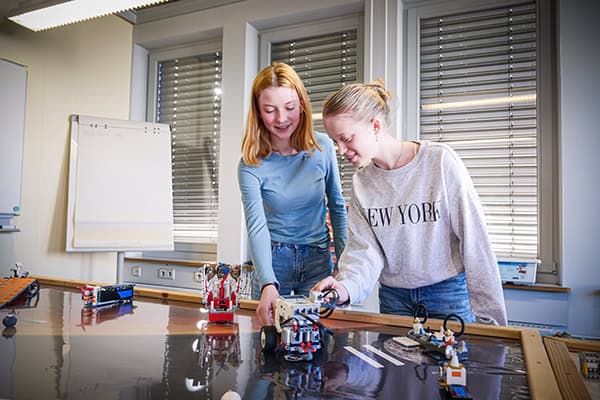 Zwei Frauen stehen an einem Tisch und gucken bzw. zeigen auf auf verschiedene LEGO-Fahrzeige, die auf dem Tisch stehen.
