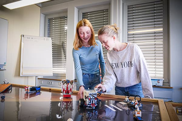 Zwei Frauen stehen an einem Tisch und gucken bzw. zeigen auf auf verschiedene LEGO-Fahrzeige, die auf dem Tisch stehen.