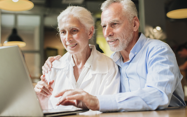 Ein älterer Mann und eine älteren Frau blicken beide auf einen aufgeklappten Laptop, der auf einem Tisch steht.