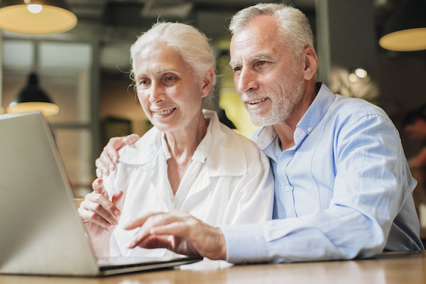 Ein älterer Mann und eine älteren Frau blicken beide auf einen aufgeklappten Laptop, der auf einem Tisch steht.