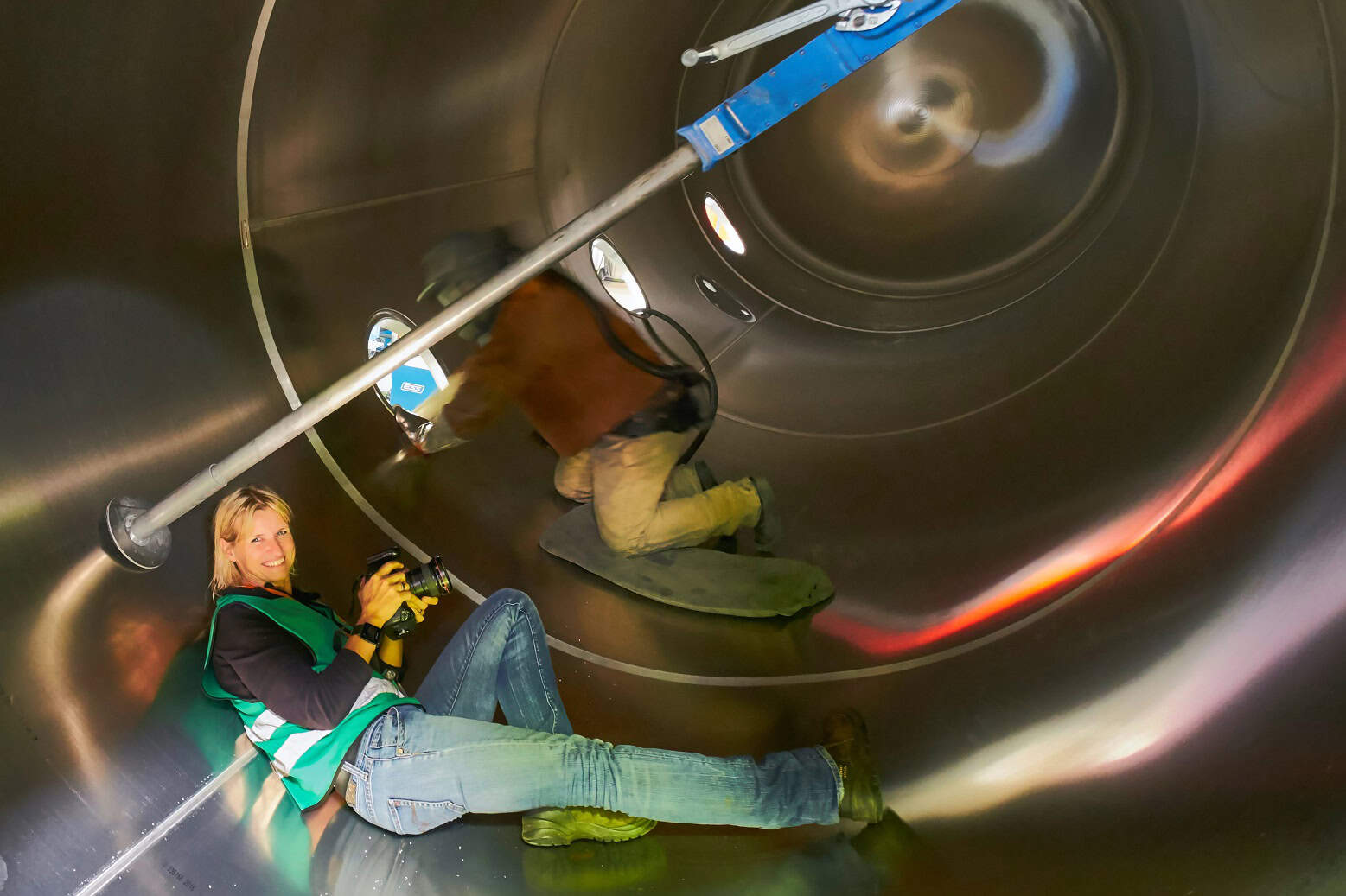 Eine Frau liegt in einer großen runden Industrie-Metallröhre , hält einen Fotoapparat in den Händen und lacht in die Kamera.