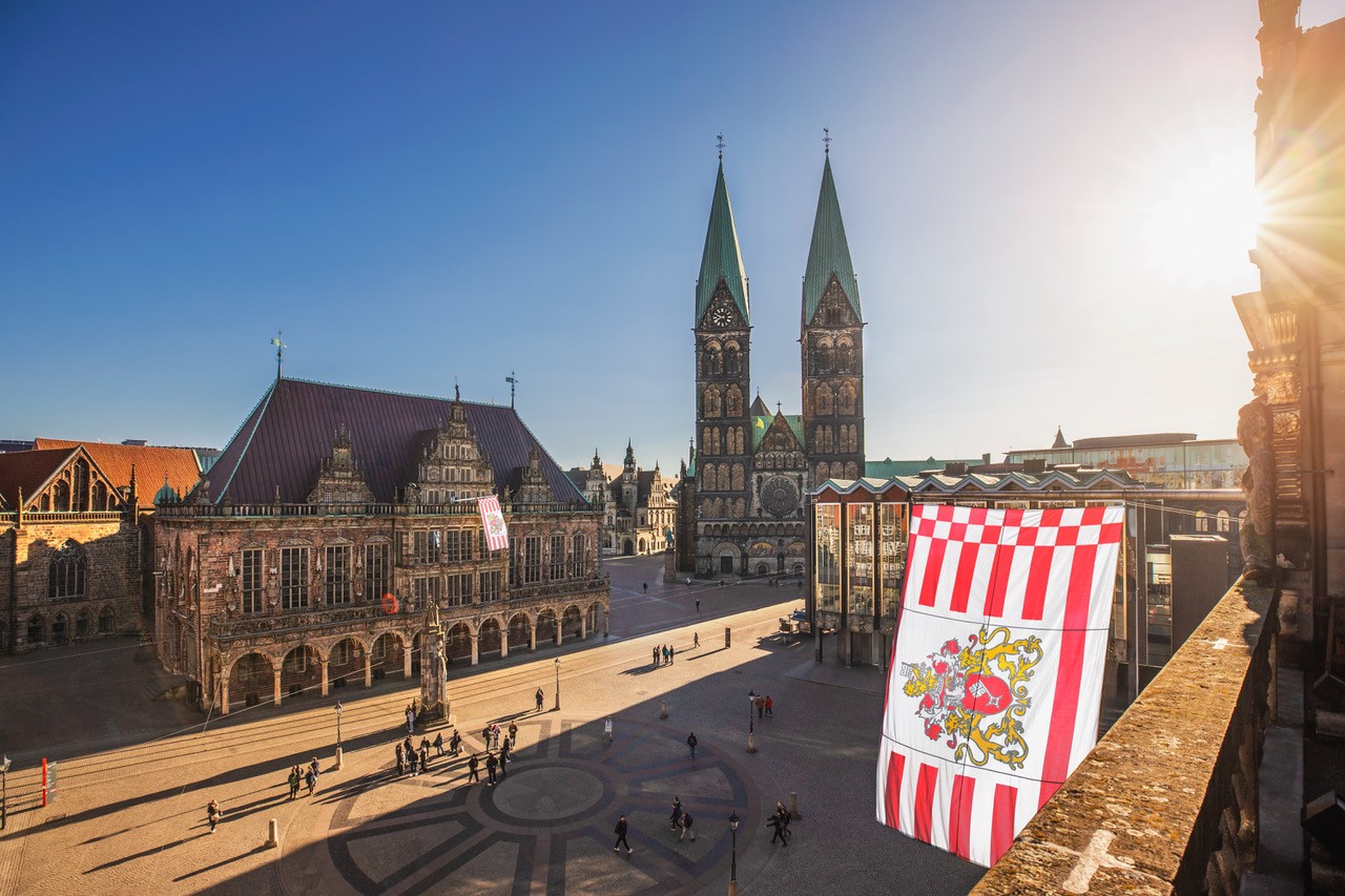 Vogelperspektive auf den Bremer Marktplatz in strahlendem Sonnenlicht. Am rechten Bildrand ist die Bremer Speckflagge zu sehen.