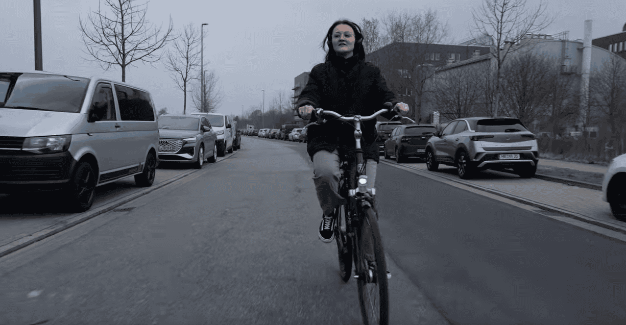 Ein Screenshot eines Videos. Eine Frau fährt auf einem Fahrrad durch eine Straße.