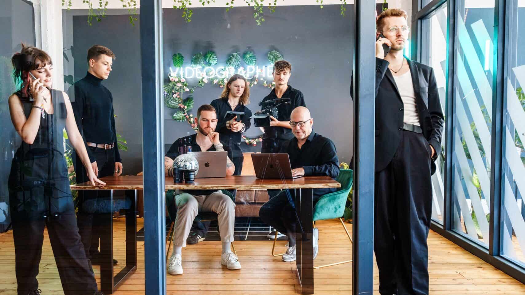 Eine Gruppe Menschen steht und sitzt hinter einer Glasfront in einem Büroraum in unterschiedlichen Arbeitsposen.