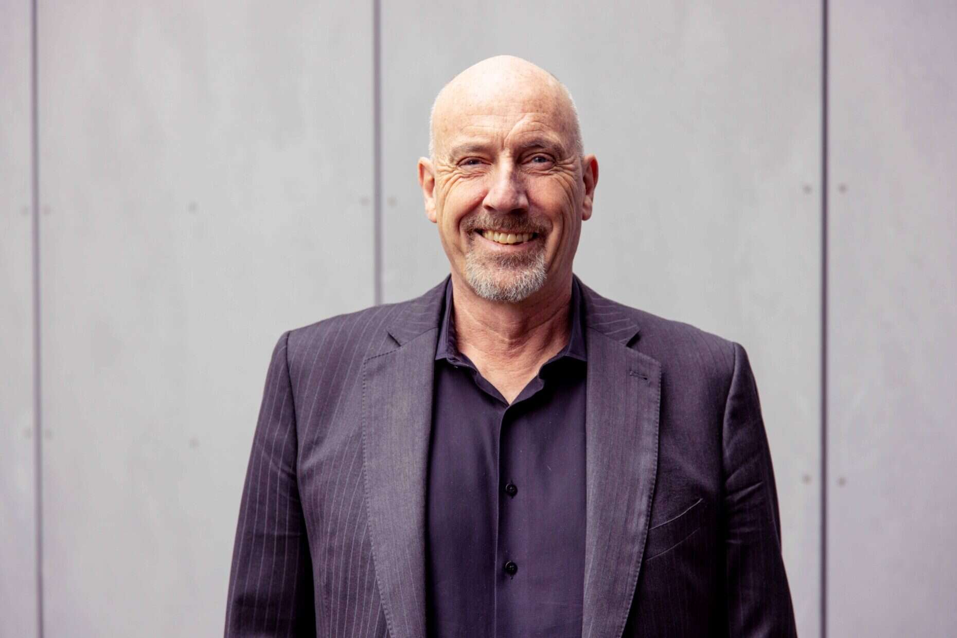 Ein Mann steht vor einer grauen Betonwadn und lächelt in die Kamera.