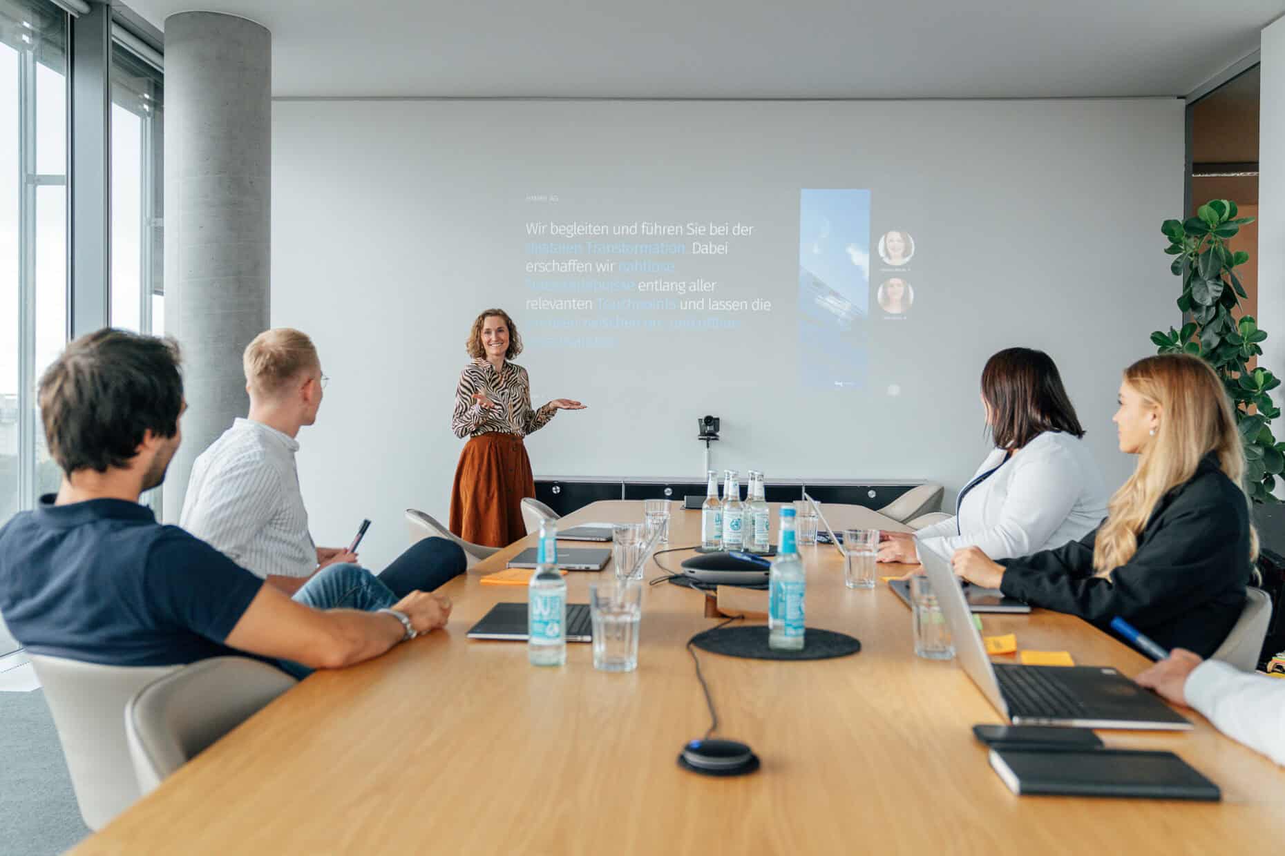 Mehrere Personen sitzen an einem Konferenztisch und schauen auf eine Frau, die vorne etwas präsentiert.