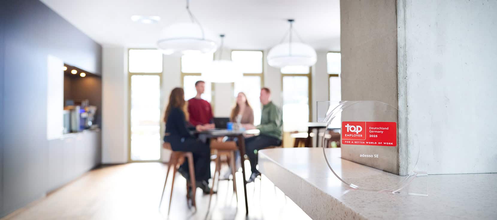 Im Hintergrund sitzen mehrere Personen an einem Tisch und sind unscharf. Im Vordergrund ist ein gläserner Preis mit der Aufschrift: Top Employer Deutschland 2025, adesso SE.