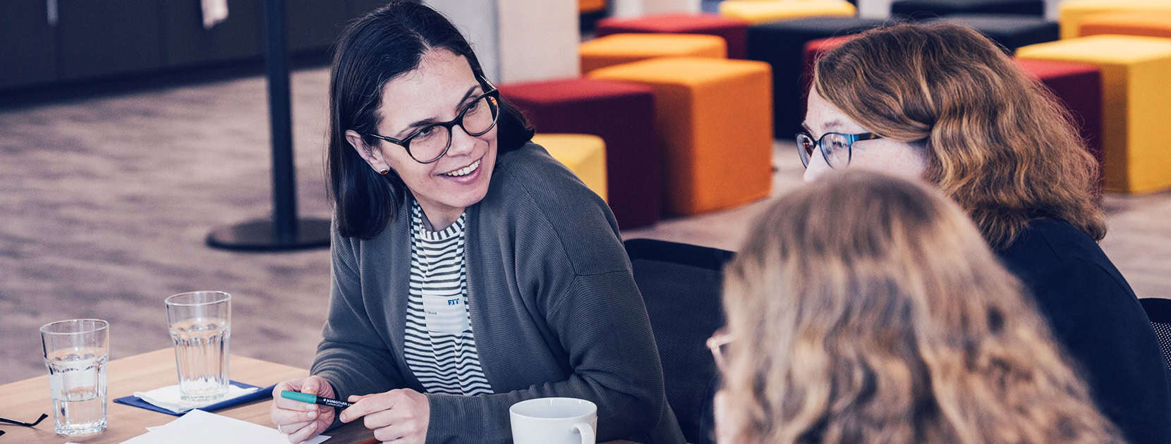 Drei Frauen sitzen an einem Tisch mit Gläsern und unterhalten sich miteinander.