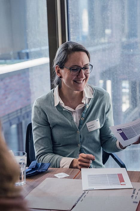 Eine Frau sitzt an einem Tisch, lächelt und hält zwei Papiere in der Hand.