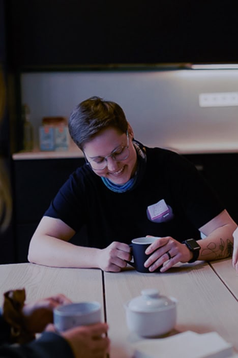 Eine Frau sitzt an einem Tisch und lacht. In ihren Händen hält sie eine Tasse.