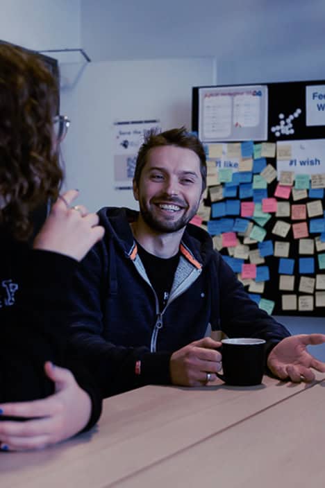 Ein Mann und eine Frau sitzen an einem Tisch und unterhalten sich. Im Hintergrund ist eine Wand mit Post-Its zu sehen.