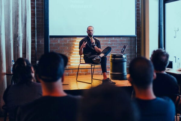 Auf einer Bühne sitzt ein Mann und spricht in ein Mikrofon. Davor sitzen mehrere Personen im Publikum.