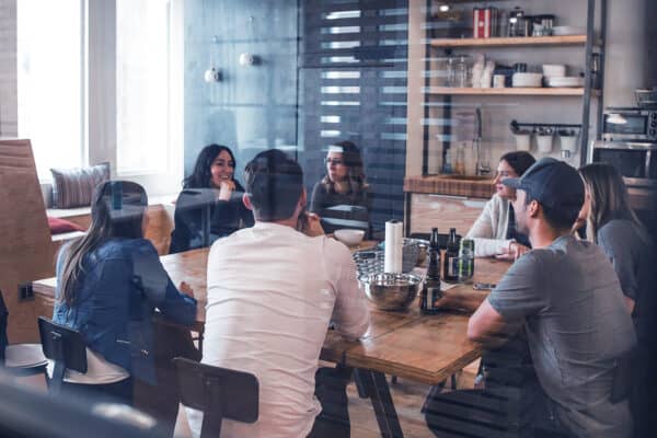 Sieben Personen sitzen an einem Tisch und sprechen miteinander. Auf dem Tisch stehen Getränke und Schüsseln.