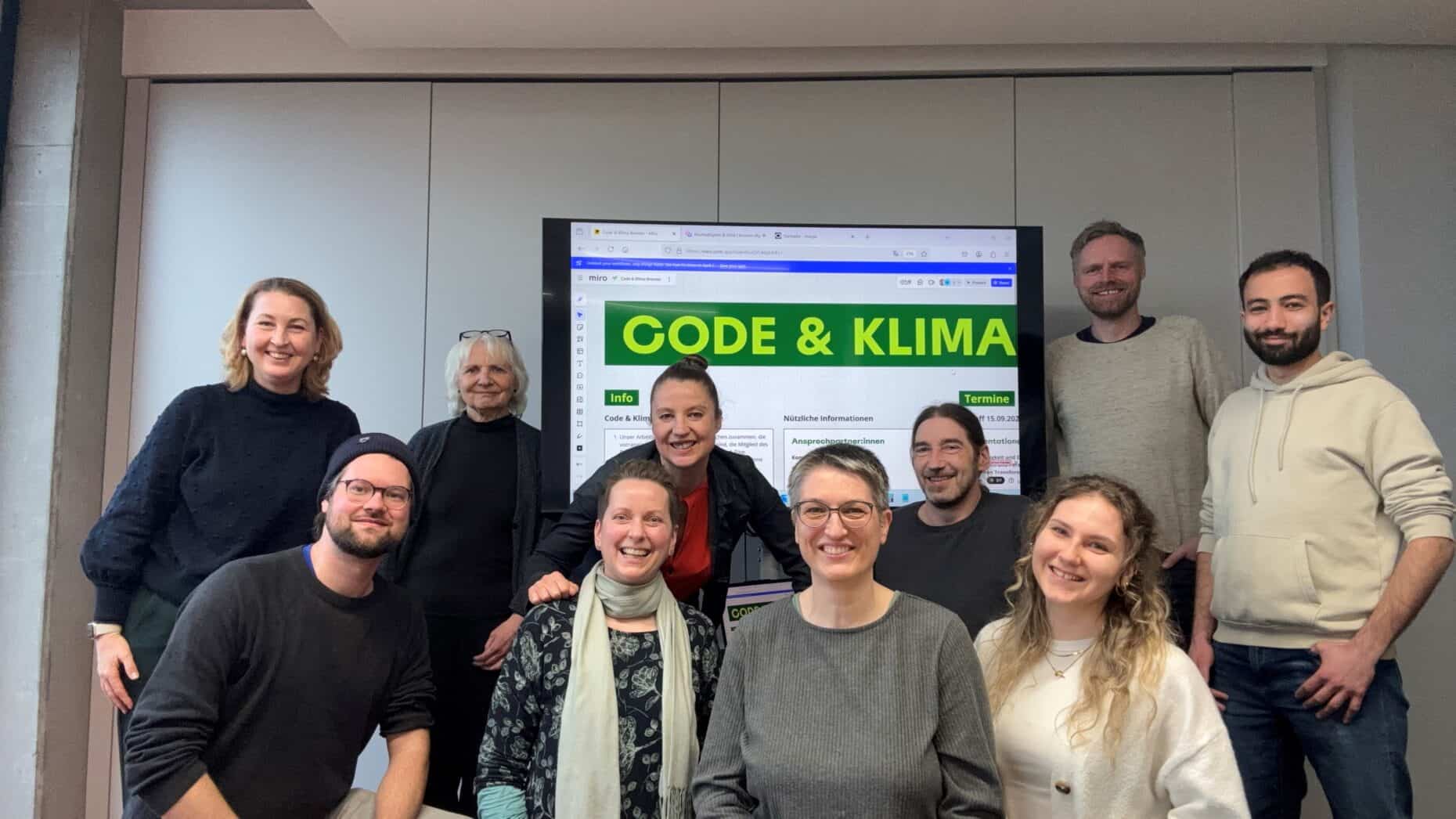Gruppe von zehn Menschen, die um einen TV-Bildschirm herum aufgestellt sind und alle in die Kamera lächeln.