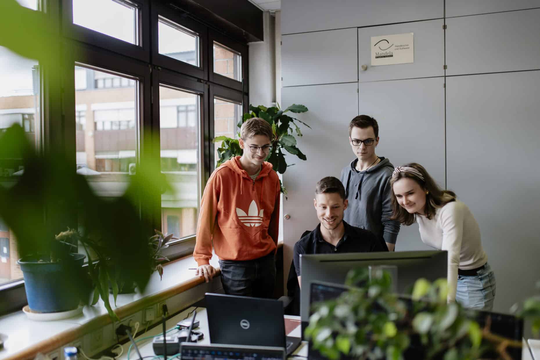 Vier Menschen sitzen und stehen inmitten eines Büros am Fenster um einen Recher auf einem Schreibtisch und schauen alle auf den Bildschirm. Im Vordergrund sind die stark verschwommenen Umrisse einer Pflanze zu erkennen.
