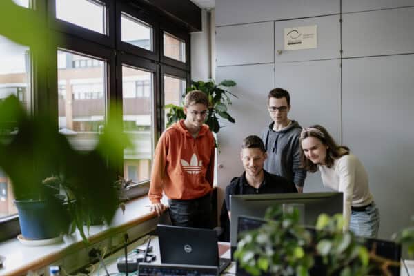 Vier Menschen sitzen und stehen inmitten eines Büros am Fenster um einen Recher auf einem Schreibtisch und schauen alle auf den Bildschirm. Im Vordergrund sind die stark verschwommenen Umrisse einer Pflanze zu erkennen.