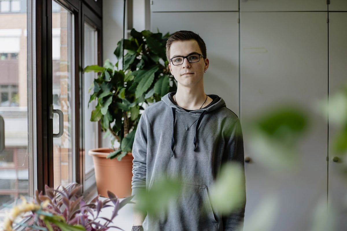 Ein Mann steht inmitten eines Büros am Fenster und schaut in die Kamera. Im Vordergrund sind die stark verschwommenen Umrisse einer Pflanze zu erkennen.