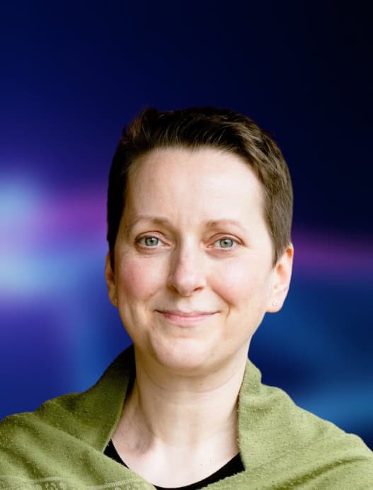 Portrait einer Person, die freundlich in die Kamera guckt. Der dunkelblau-liafarben und verschwommene Hintergrund ist in der Bildberarbeitung nachträglich hinzugefügt worden.