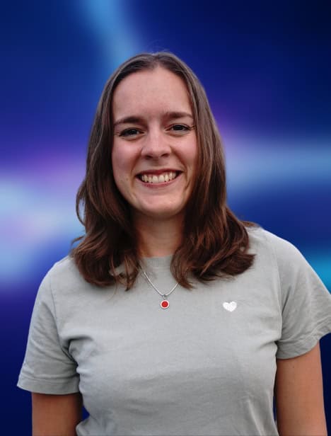 Portrait einer Person, die freundlich in die Kamera guckt. Der dunkelblau-liafarben und verschwommene Hintergrund ist in der Bildberarbeitung nachträglich hinzugefügt worden.