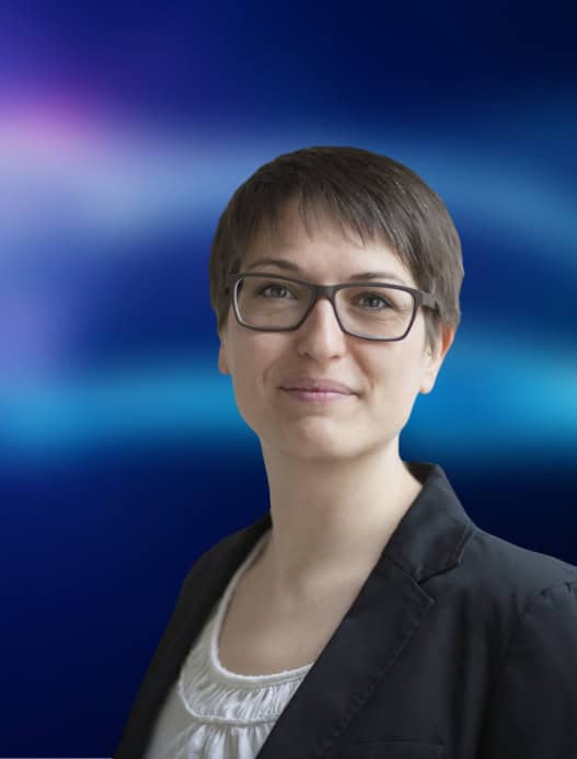 Portrait einer Person, die freundlich in die Kamera guckt. Der dunkelblau-liafarben und verschwommene Hintergrund ist in der Bildberarbeitung nachträglich hinzugefügt worden.