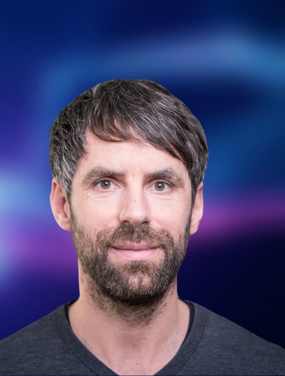 Portrait einer Person, die freundlich in die Kamera guckt. Der dunkelblau-liafarben und verschwommene Hintergrund ist in der Bildberarbeitung nachträglich hinzugefügt worden.