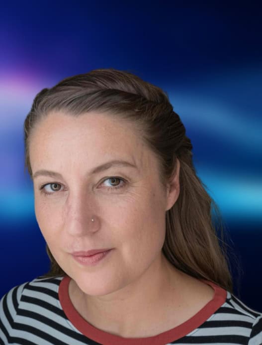 Portrait einer Person, die freundlich in die Kamera guckt. Der dunkelblau-liafarben und verschwommene Hintergrund ist in der Bildberarbeitung nachträglich hinzugefügt worden.