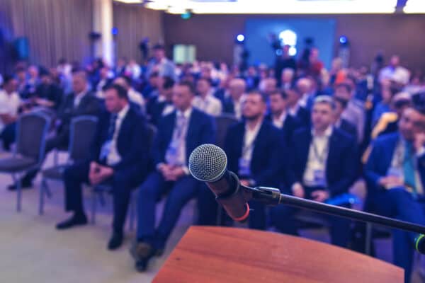 Nahaufnahme eines Microfons im Ständer. Im Hintergrund sind verschwommen sitzende Menschen in Stuhlreihen zu erkennen.