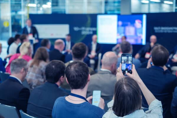 Blick in eine Talkrunde auf einer Messe oder Konferenz. Im vorderen Bildbereich sitzen Menschen mit dem Rücken zum Foto und blicken auf die Menschen, die sitzend verschwommen zu erkennen sind.