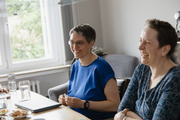 Zwei Frauen sitzen lachend an einem Tisch und schauen aus dem Foto wahrscheinlich auf Personen gegenüber des Tisches.
