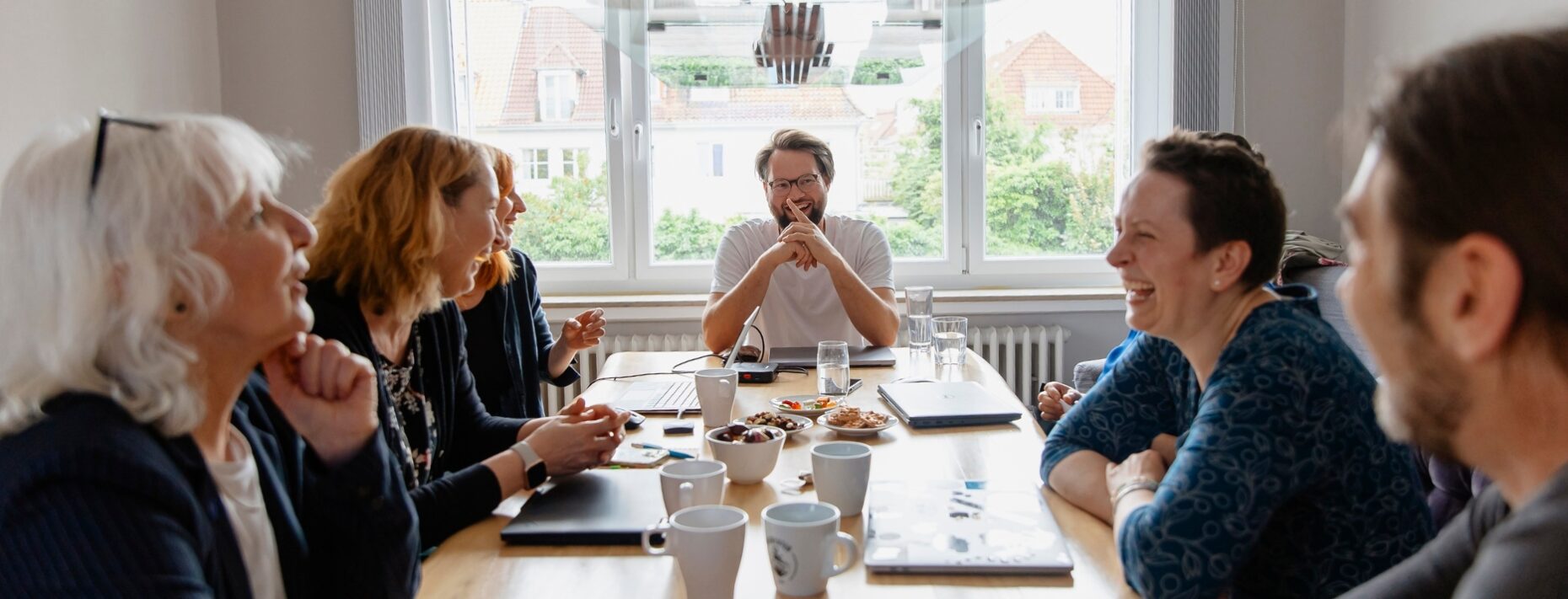 Foto einer Gruppe lachender Menschen, die alle um einen großen Holztisch herum sitzen.