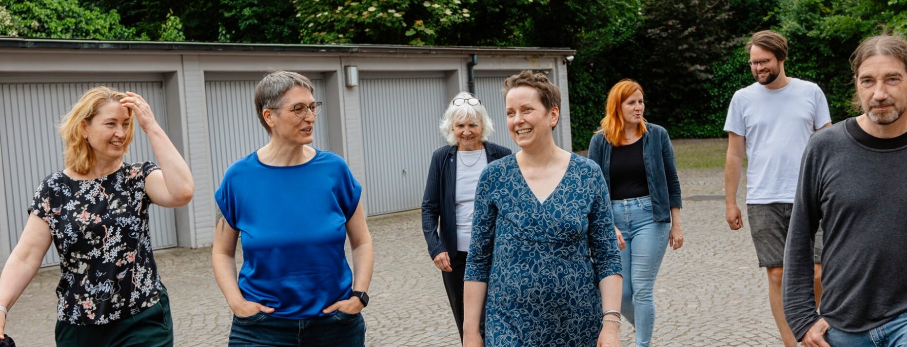 Anschnitt einer Gruppe von Menschen, die sich unterhaltend draußen über einen Garagenhof gehen und auf den/die Betrachter*in zugehen.