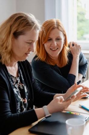 Zwei Frauen sitzen an einem Holztisch. Die eine hat ein Smartphone in der Hand und schaut darauf, die andere schaut über die Schulter mit aufs Smartphone.