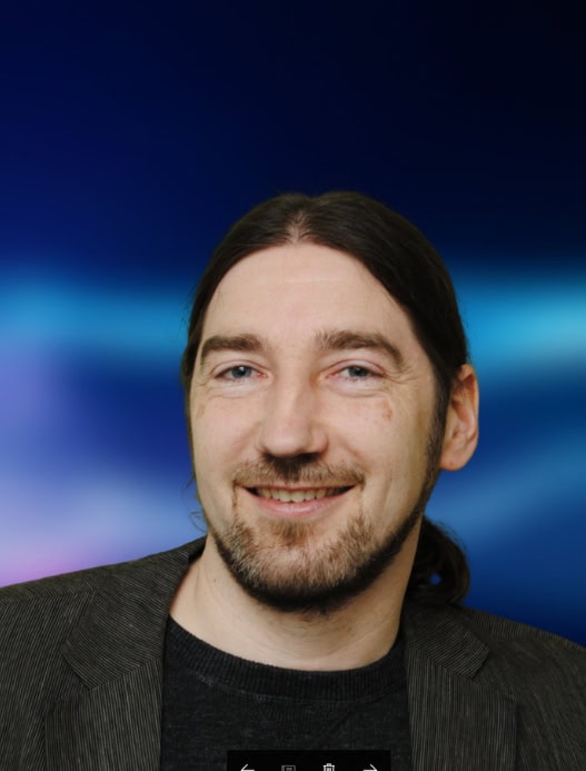 Portrait einer Person, die freundlich in die Kamera guckt. Der dunkelblau-liafarben und verschwommene Hintergrund ist in der Bildberarbeitung nachträglich hinzugefügt worden.