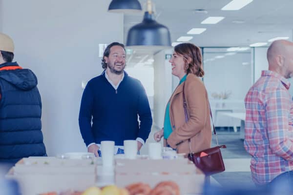 Zwei Menschen stehen in einem hellen Raum am Ende eines Tisches miteinader im Gespräch und lachen.