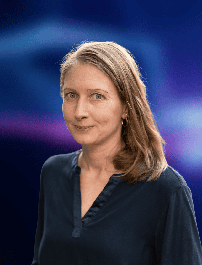 Portrait einer Person, die freundlich in die Kamera guckt. Der dunkelblau-liafarben und verschwommene Hintergrund ist in der Bildberarbeitung nachträglich hinzugefügt worden.