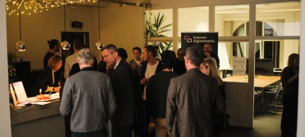 Blick in einen Innenraum mit Glasfronten und einem Tresen an dem bei gedimmter Beleuchtung verschiedene Menschen zusammenstehen und in Gespräche vertieft sind.